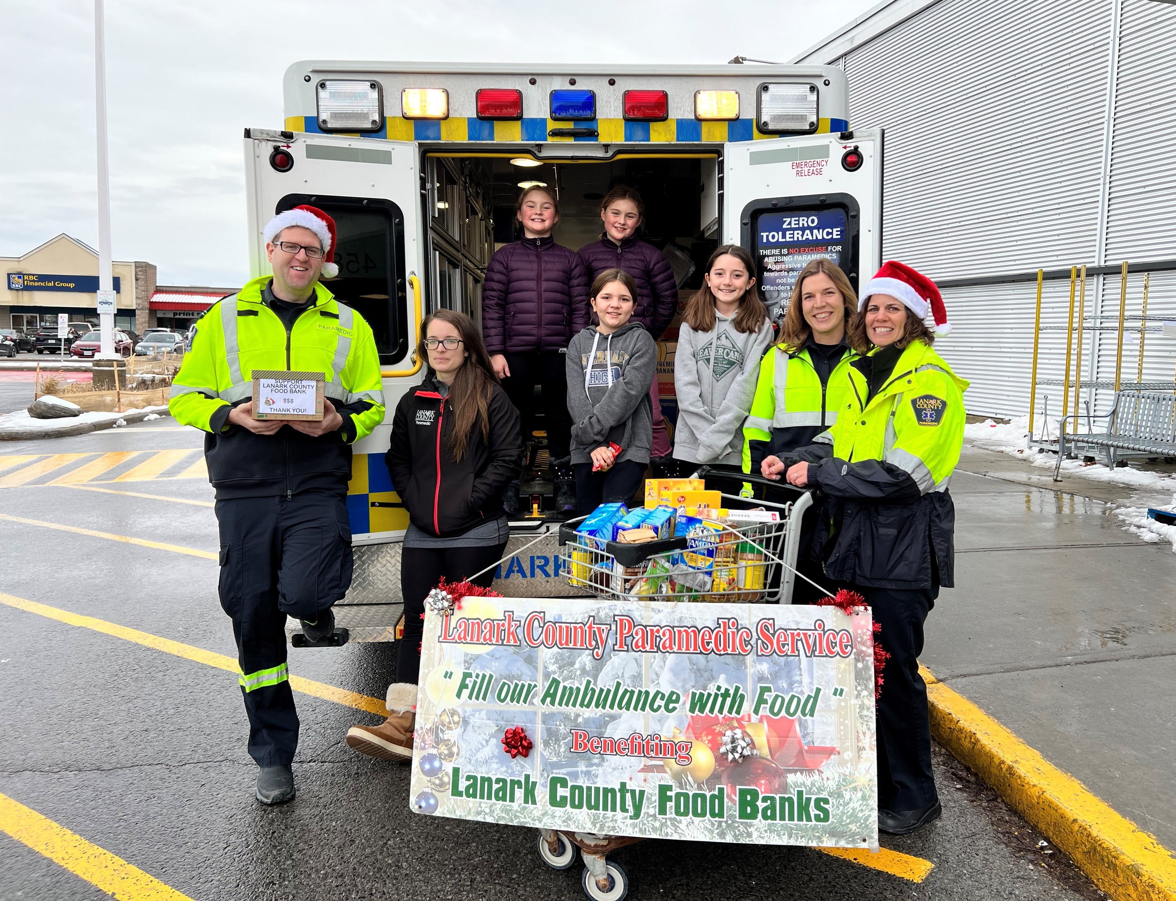 Paramedic service members and volunteers stand at the back of an ambulance with a food drive sign