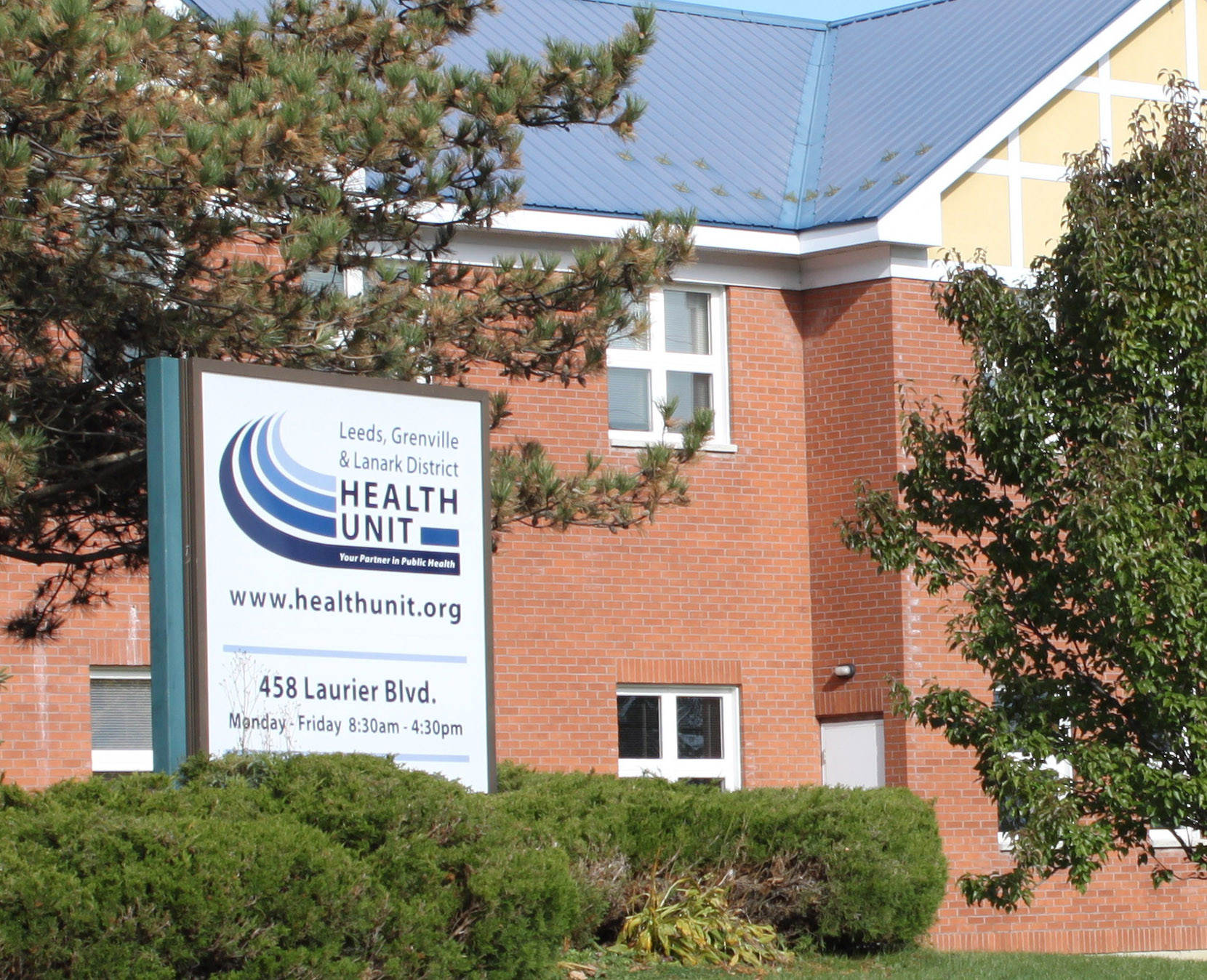 A blue and white sign advertising the Leeds, Grenville and Lanark District Health Unit sits in front of a two-storey red brick building with trees surrounding it