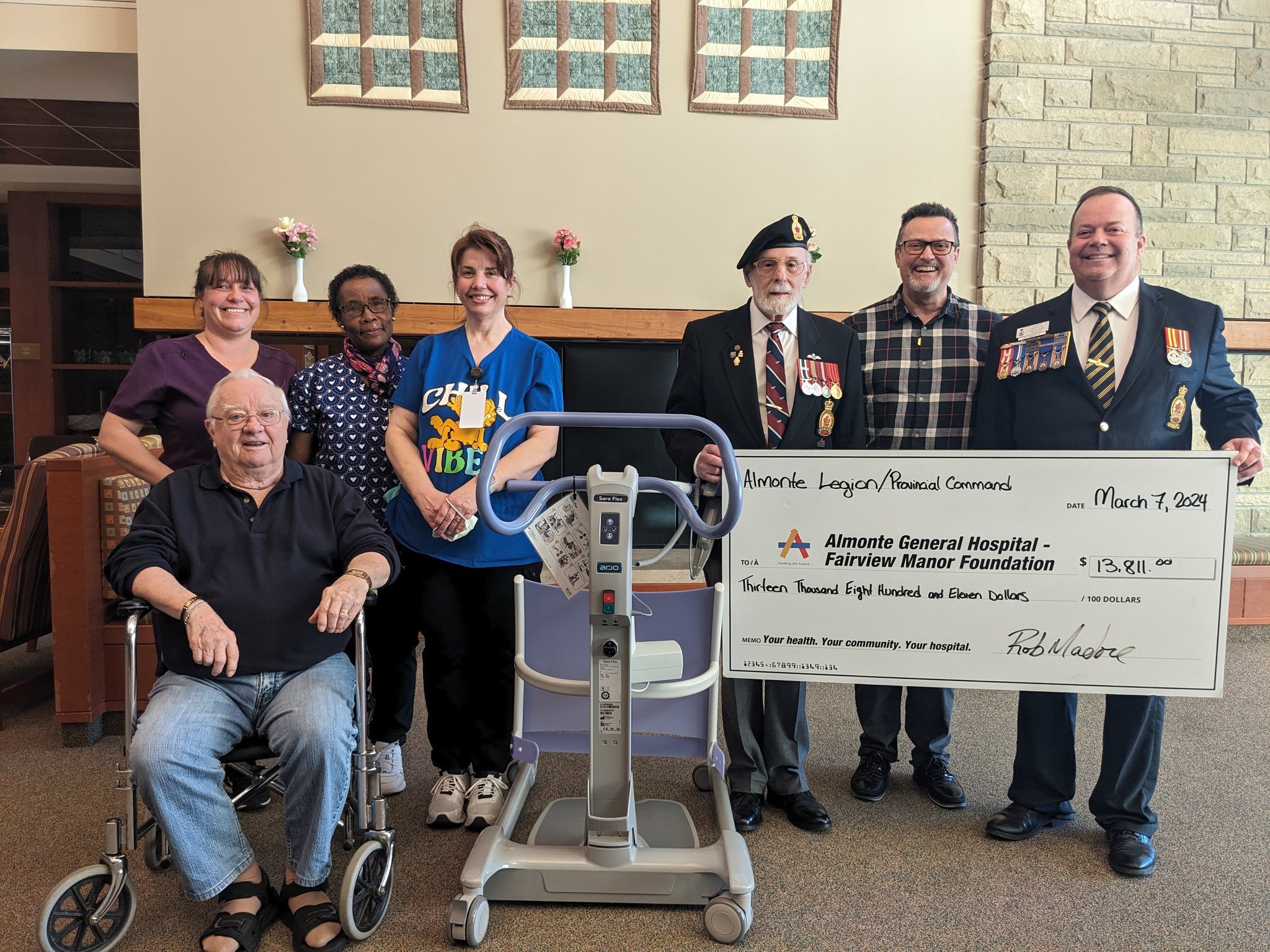 Seven people group together for a photo with one man to the left in a wheelchair. Three men on the right hold a large cheque. They are surrounded by a Sara Flex Active Lift.