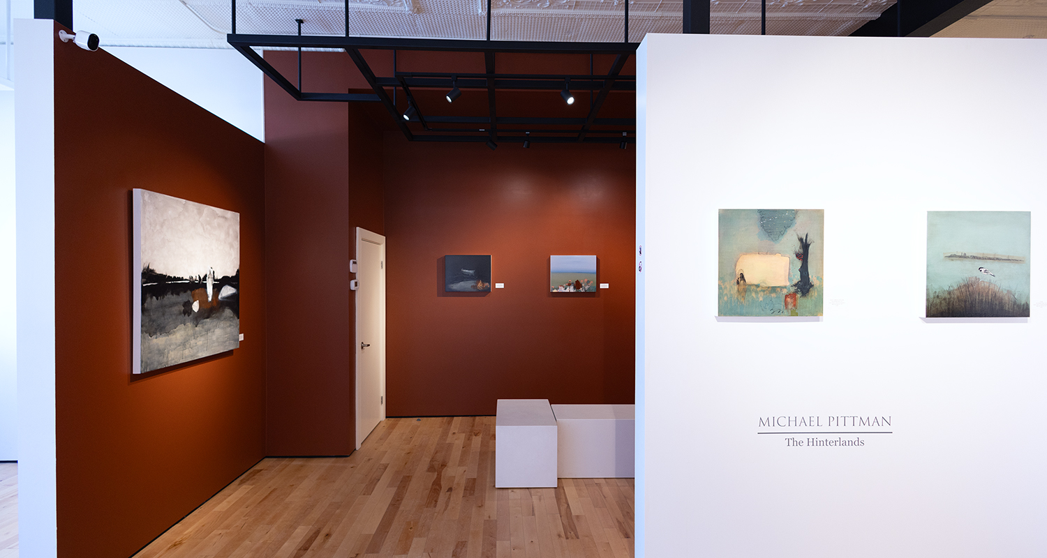 Paintings hang on brown and white gallery walls, displaying a sign reading 'Michael Pittman: The Hinterlands'