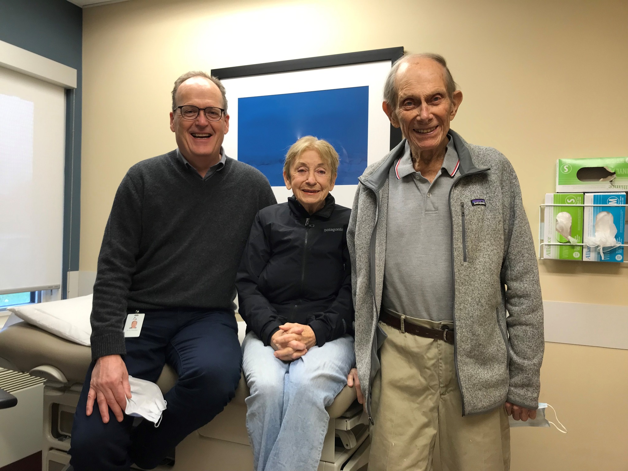 Three people smile for a photo in a doctor's exam room. Far left is a man with glasses and a grey sweater, in the middle is a woman with short blonde hair and on the right is a balding older man with a grey shirt.