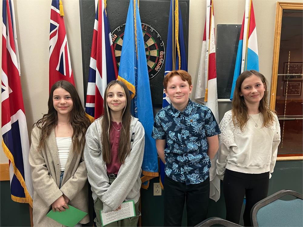 Five students (three girls and one boy) stand in front of flags in a building