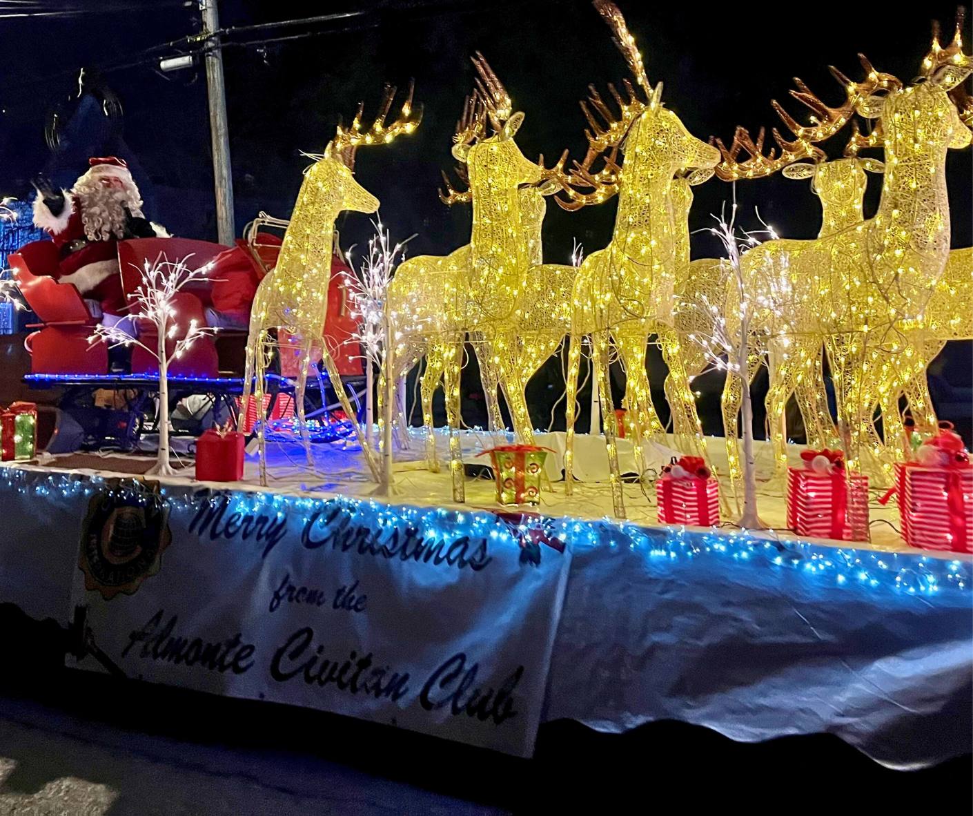 Santa Claus sits atop a sleigh in a parade float, surrounded by lighted presents and reindeer