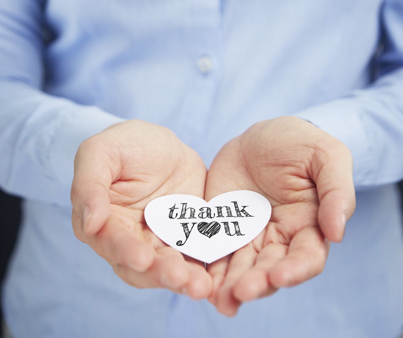 Women wearing light blue long-sleeved shirt holds palms out with a white heart-shaped note bearing the words 'thank you'