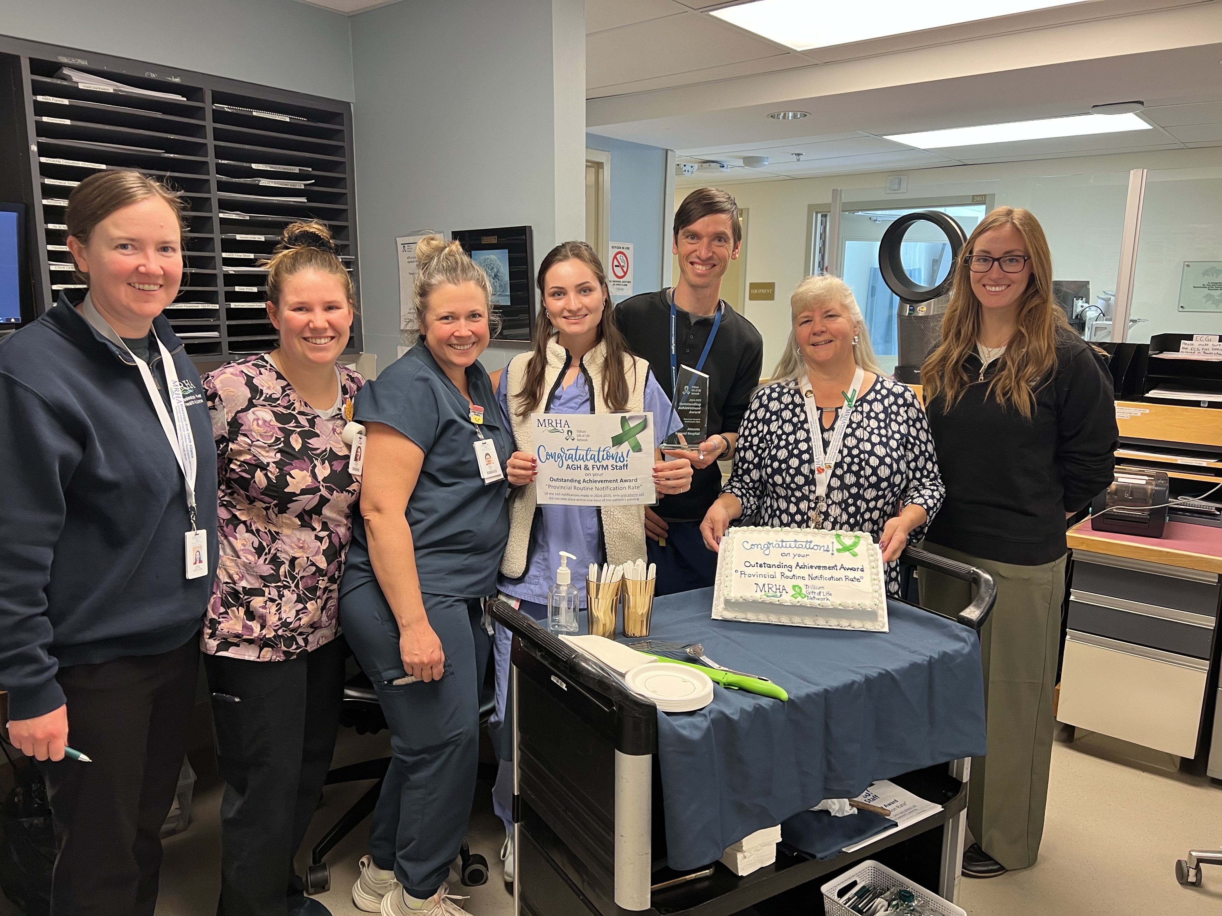 Photo of people in a hospital admin area standing for a photo. Two people hold up a sign and cake.