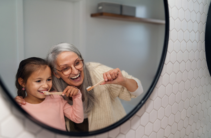 An older woman and young child stand in front of the mirror smiling as they prepare to brush their teeth