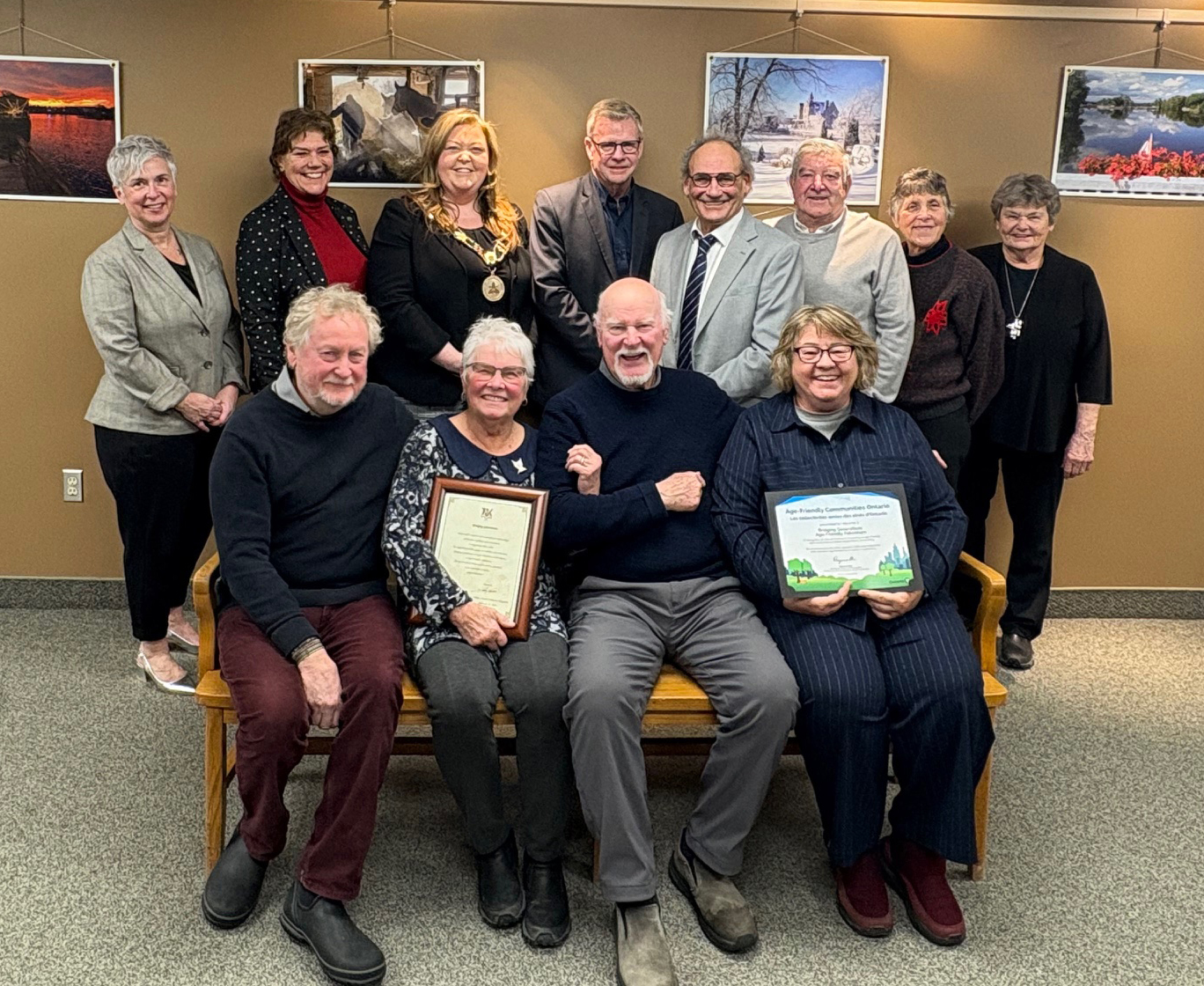 A group of people pose for a photo against a tan wall. Eight people are standing in behind four people sitting on a bench. Two people sitting hold framed certificates.
