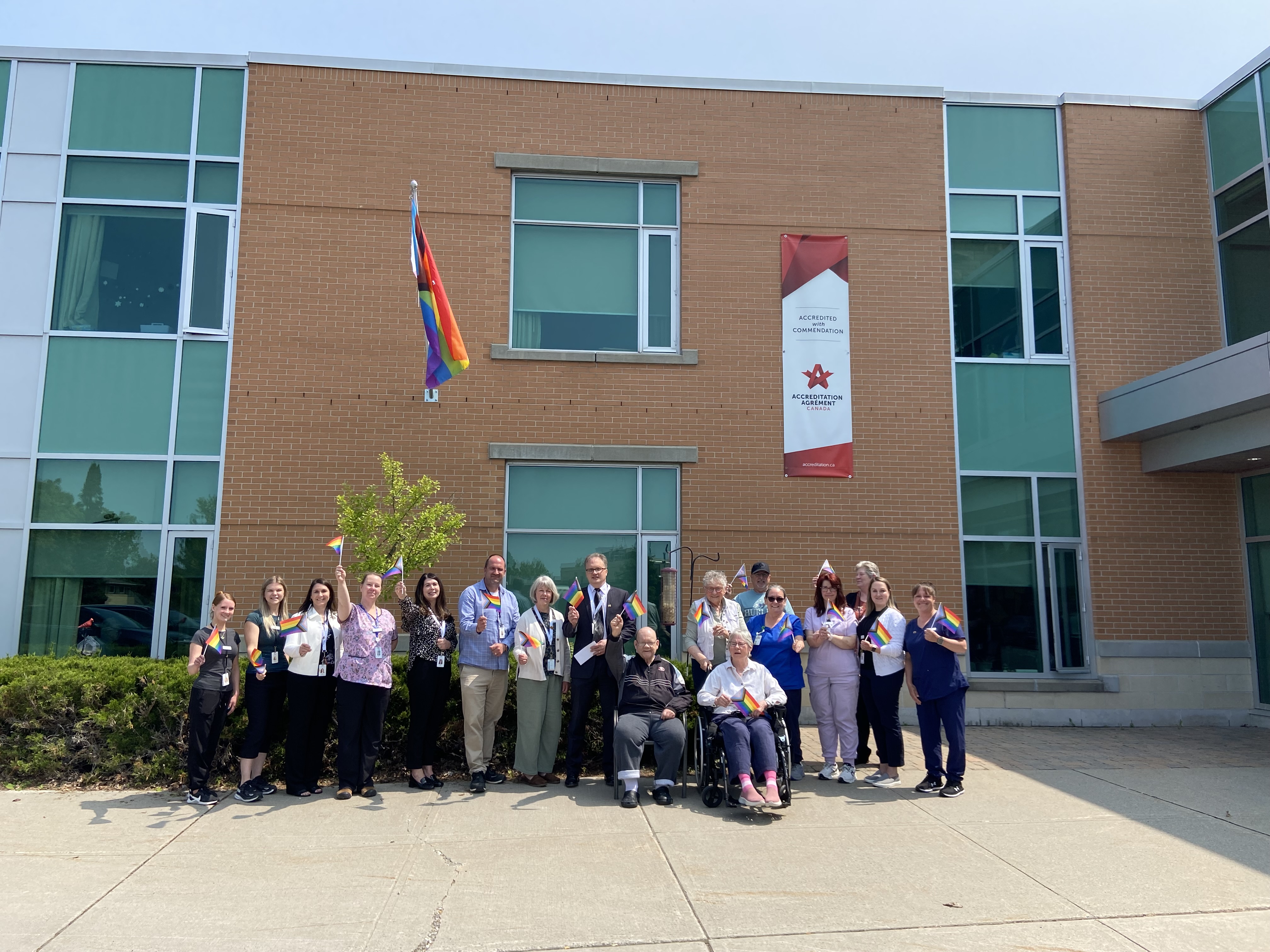 Group of people gather for a photo in front of a two-storey brick building with a Pride flag flying above.