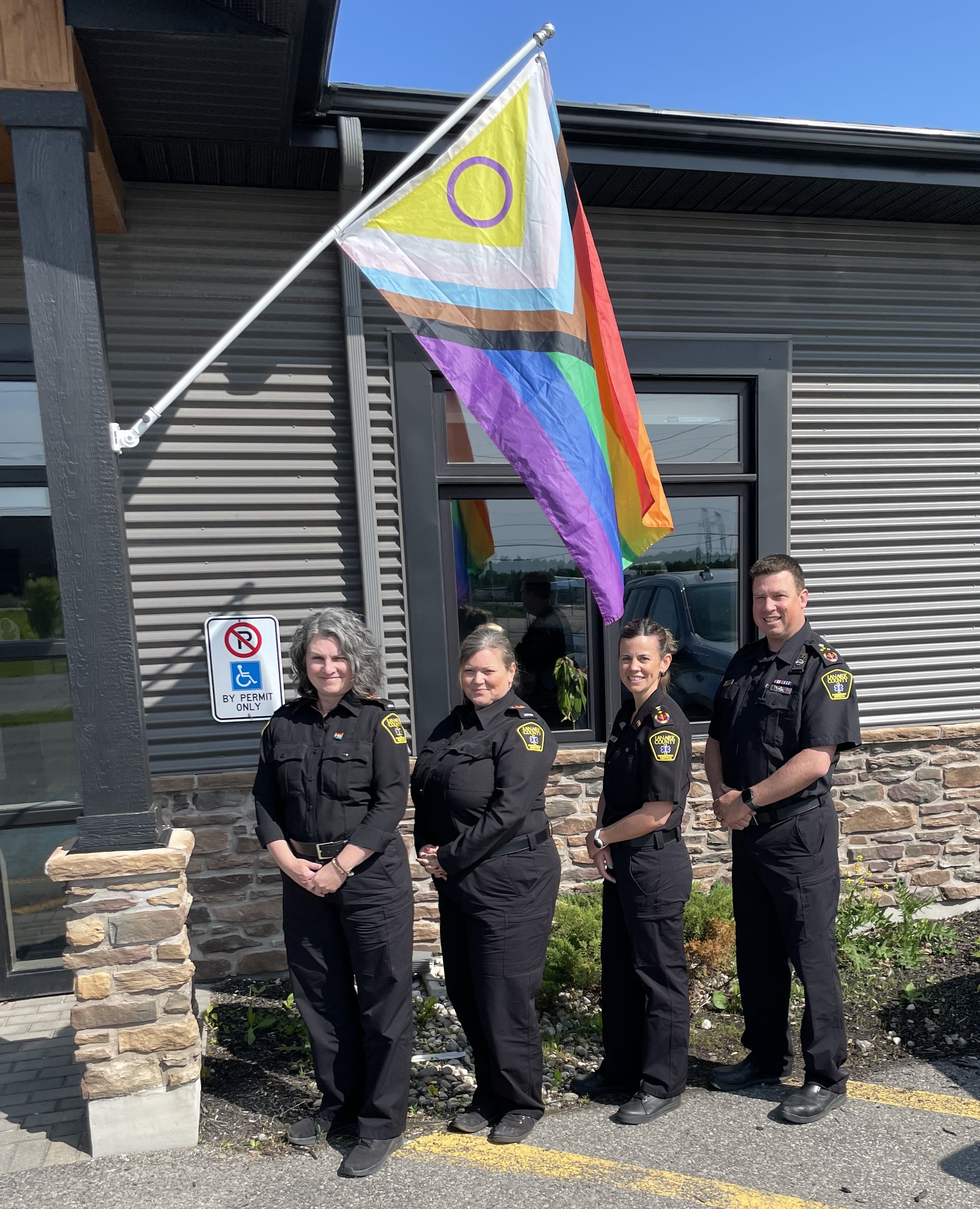 Four people in paramedic uniforms stand in front of a grey sided building underneath a Progress Pride flag