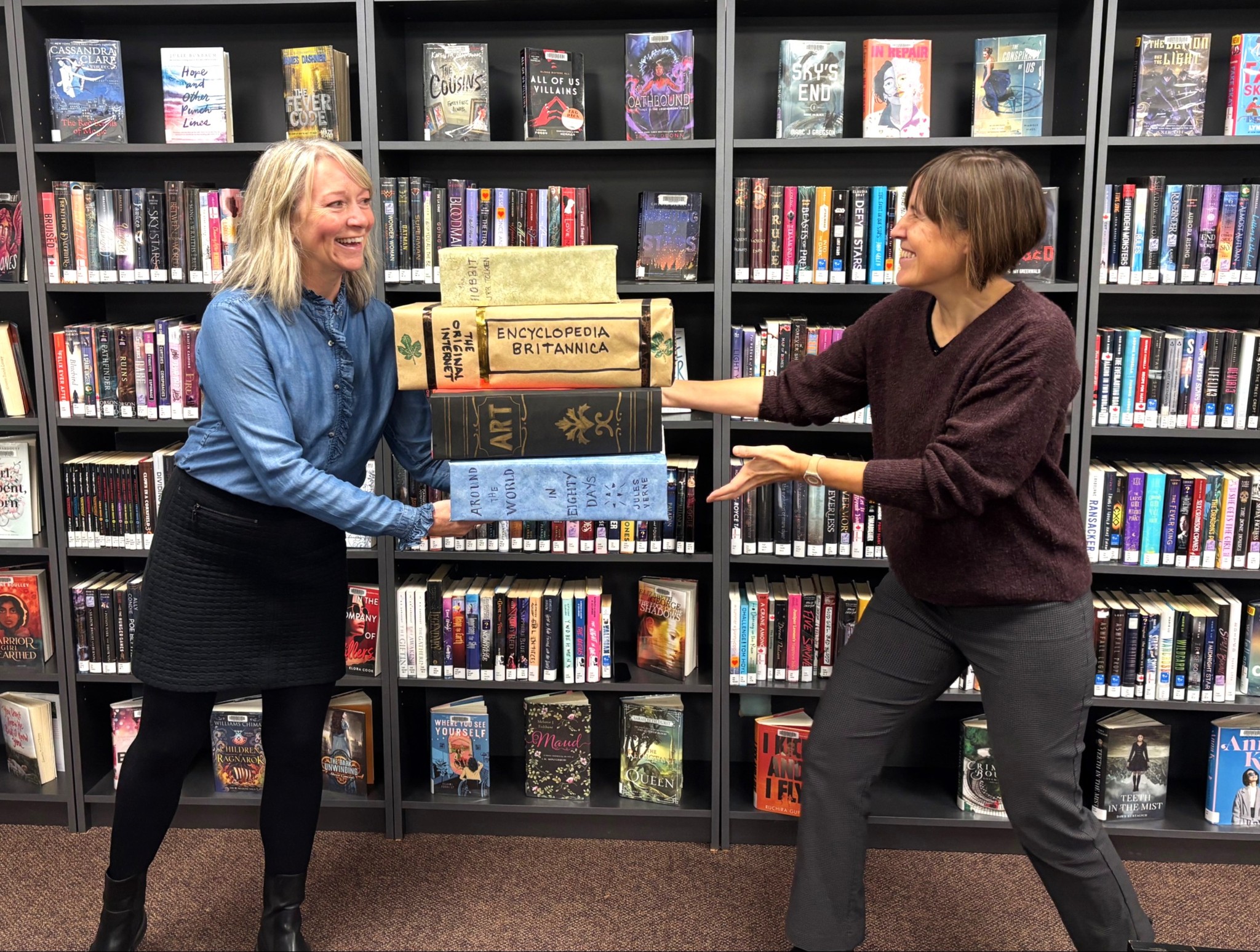 Two women stand in front of a row of bookshelves filled with books, while holding giant books.