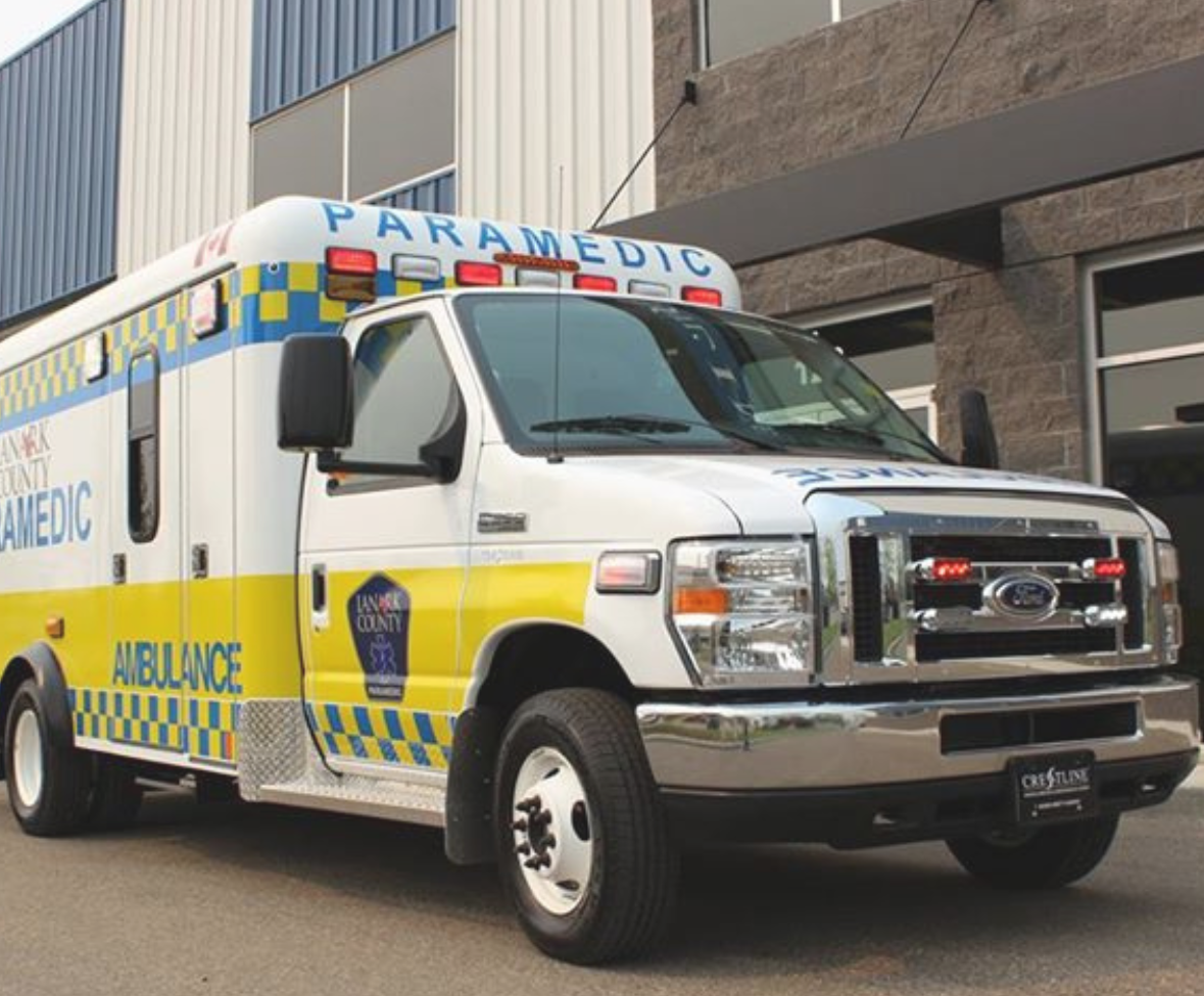 An ambulance sits parked outside of a hospital. It has blue and yellow flashing on it and reads 'Lanark County Paramedic'