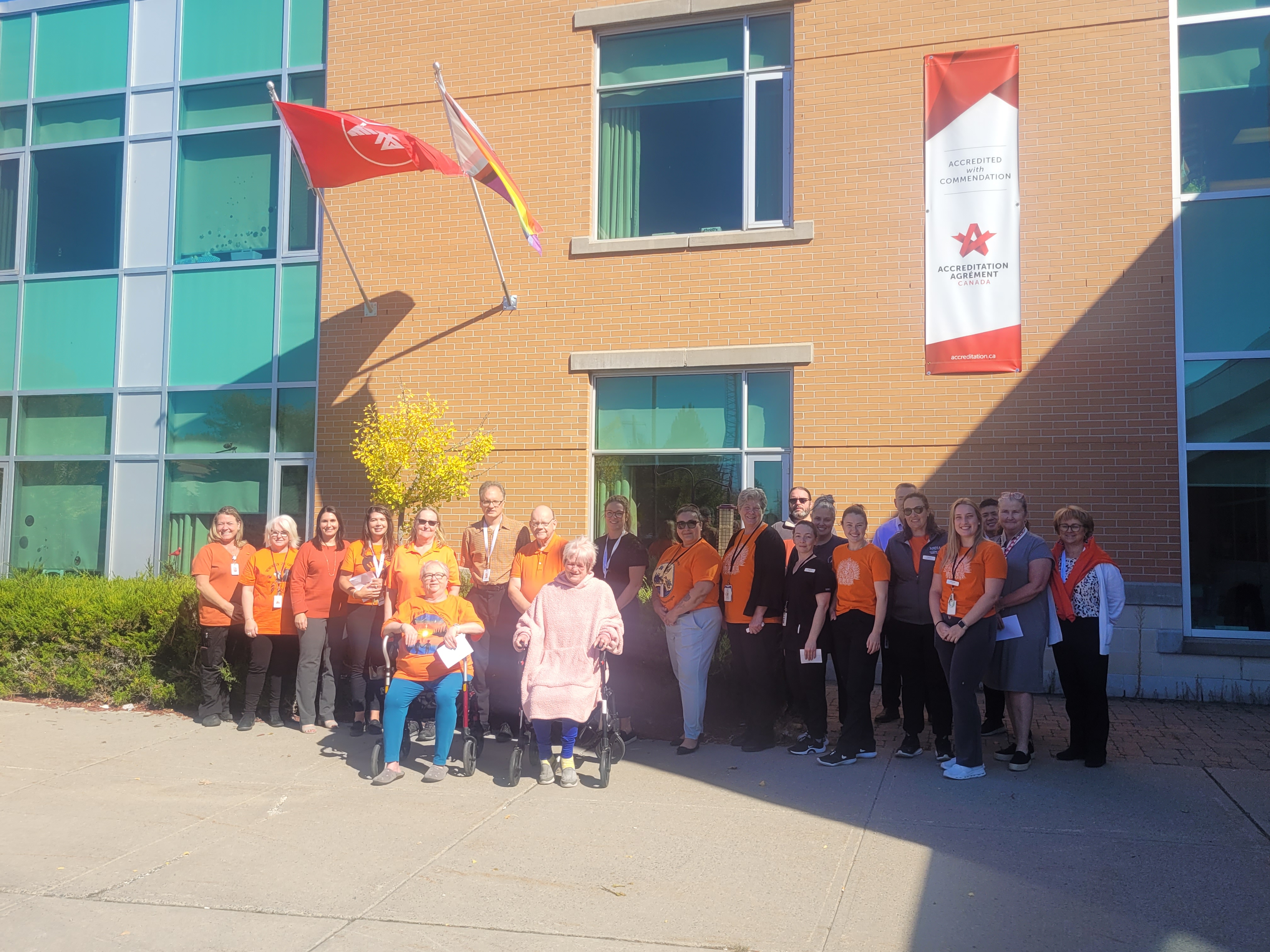 A group of people, most wearing orange shirts, gather for a photo by a building underneath flags