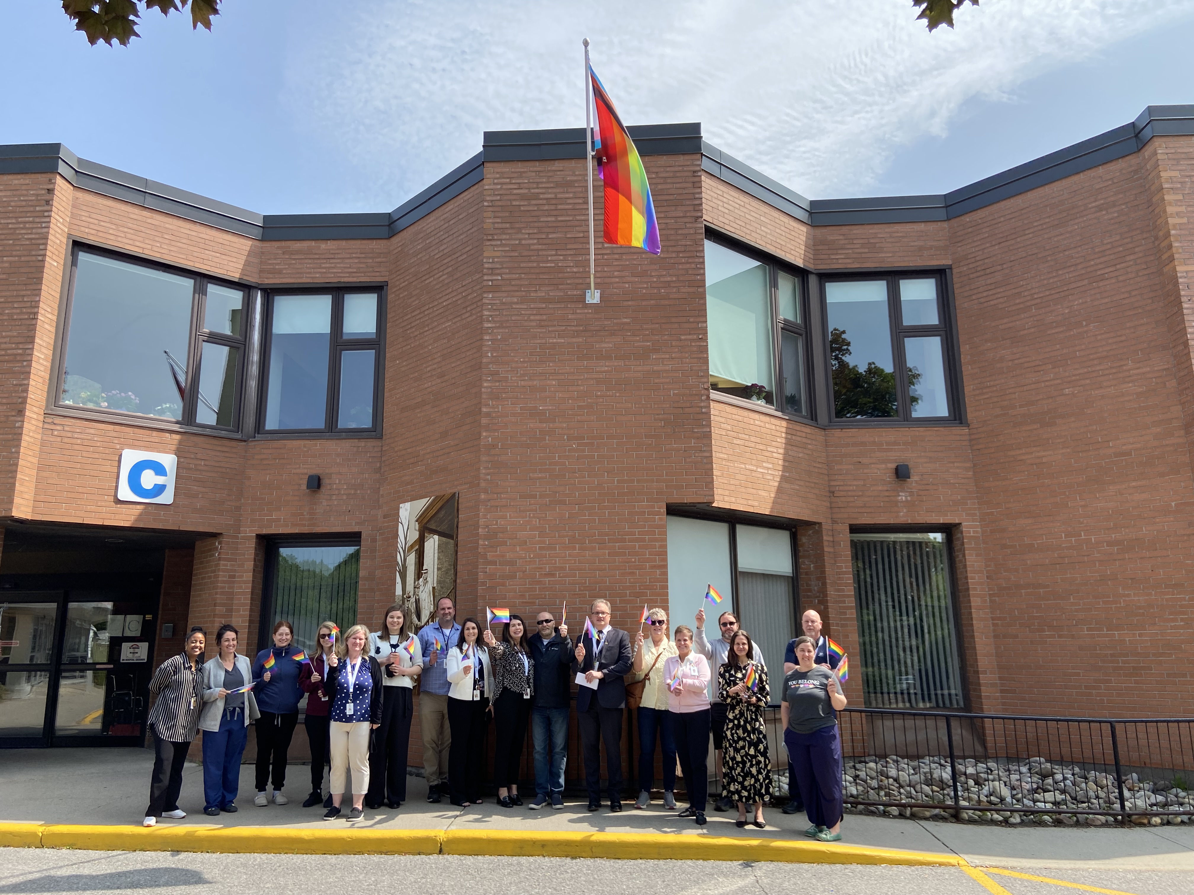 Group of people stand underneath a Pride flag flying on a red brick building