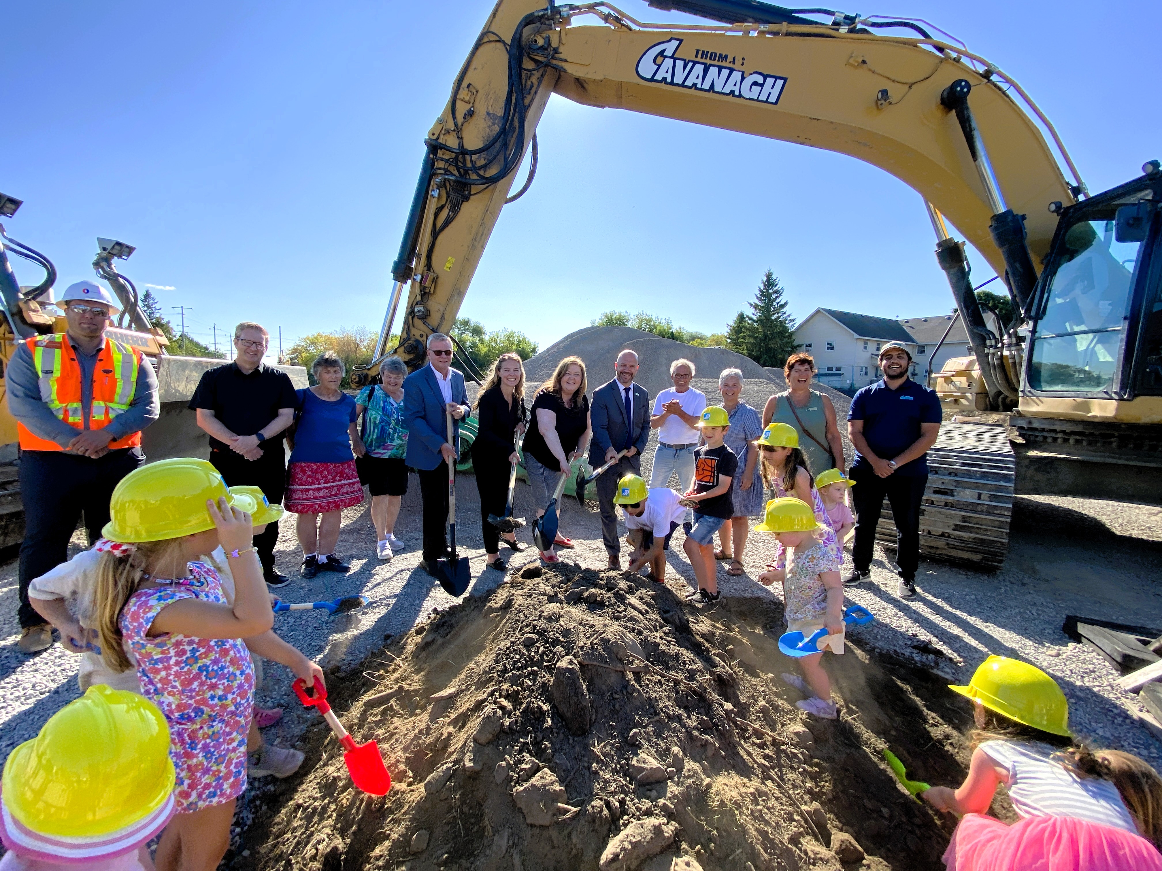 A group of people stand in front of construction equipment with shovels in a pile of dirt. Children surround pile of dirt with toy shovels.