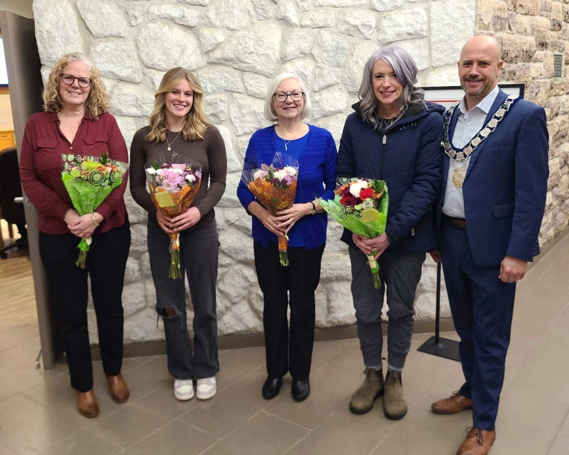 Five people stand for a photo in front of a stone wall. The four women in the photo are holding flowers.
