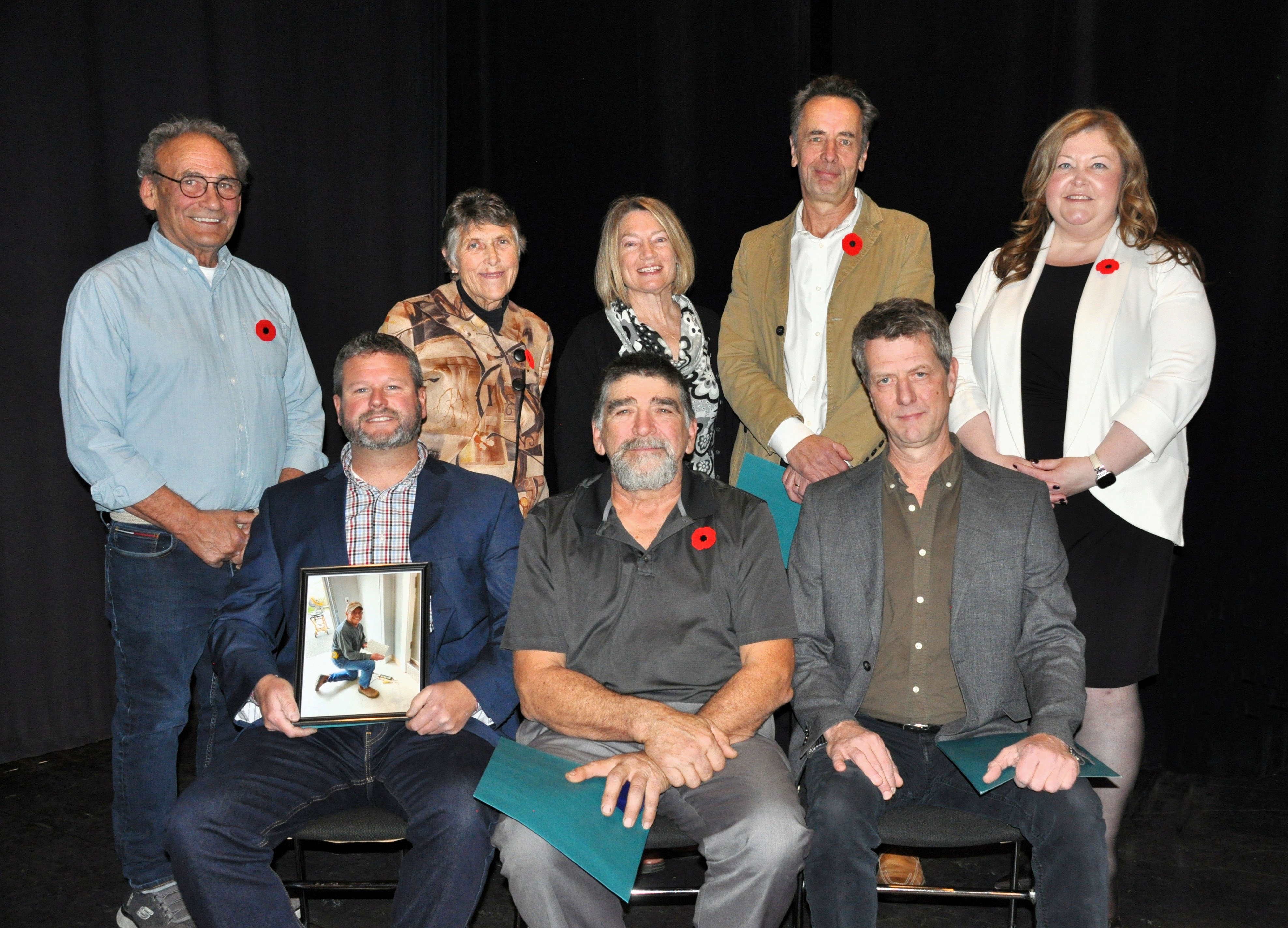 Group of people pose for a photo against a black curtain. Back row is standing, front row is seated in chairs.
