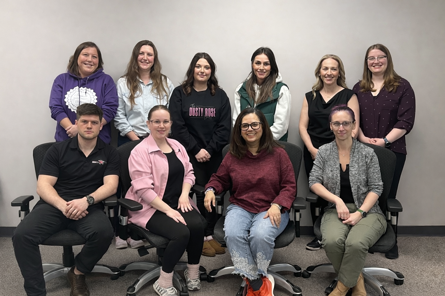 Photo of two rows of people posing for a photo against a grey wall. The back row is standing while the front row is seated.