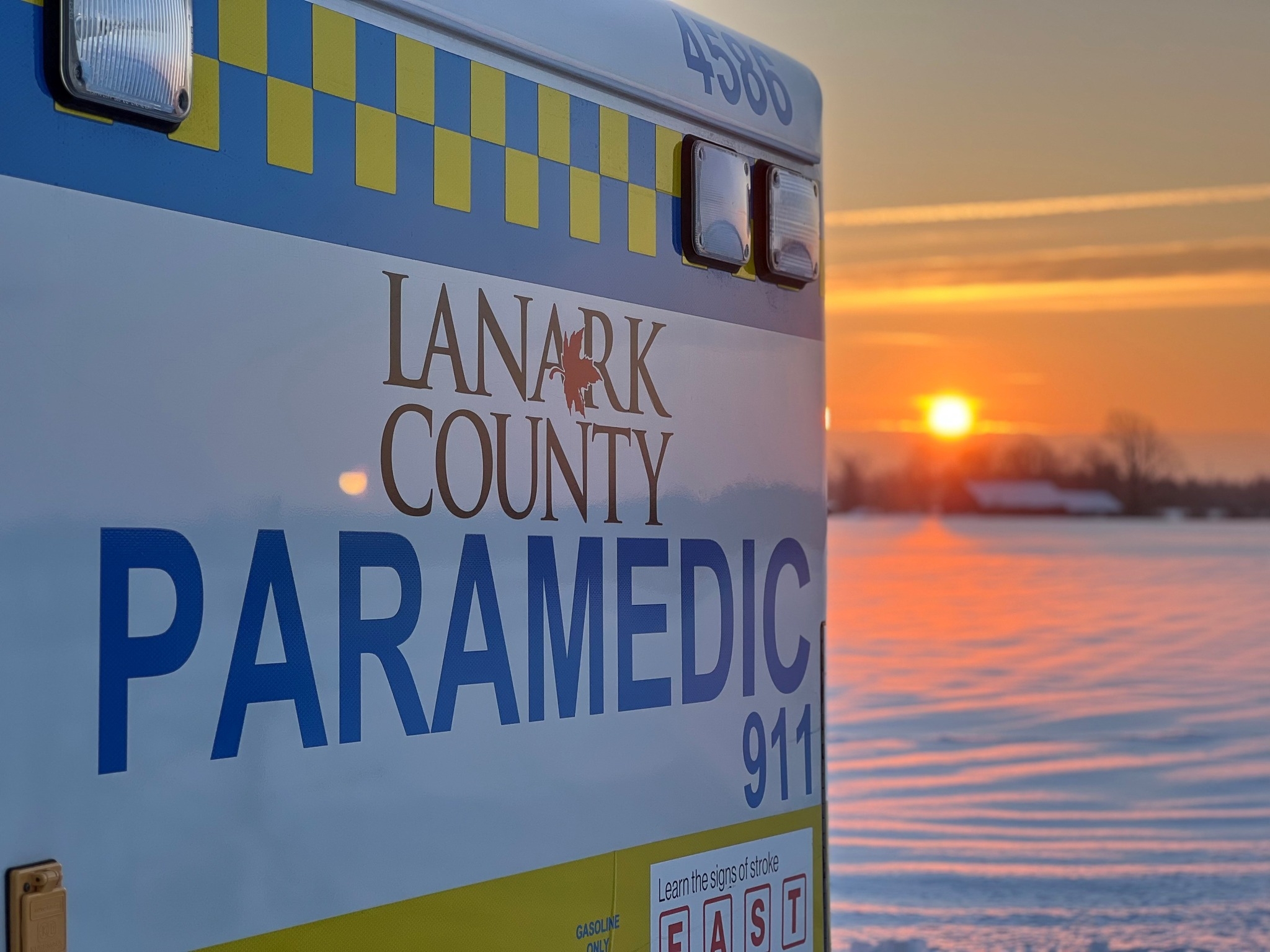 A closeup of a Lanark County Paramedic Service ambulance while the sun sets on a snowy field