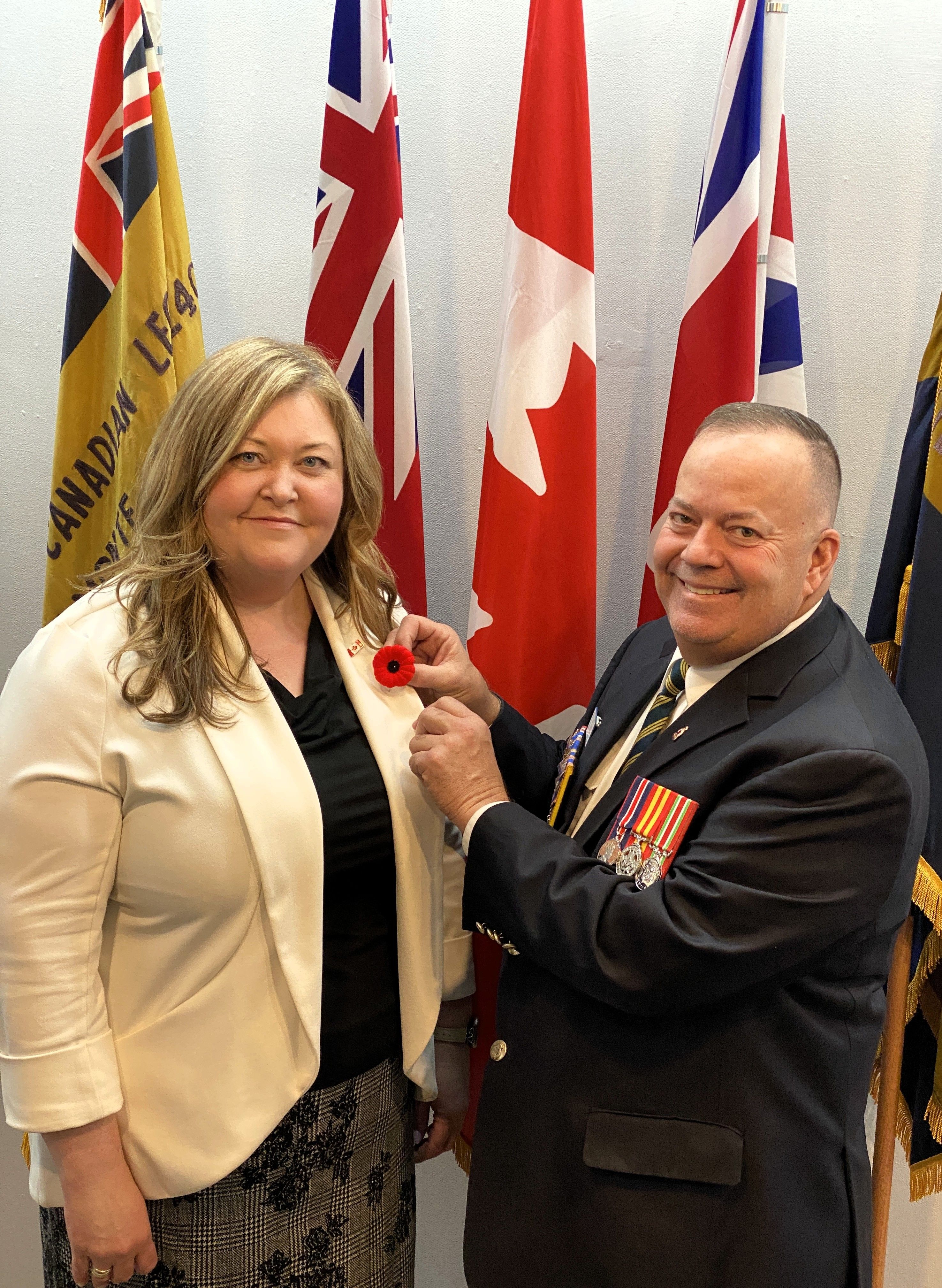Man in suit pins poppy to woman in white blazer with flags in the background