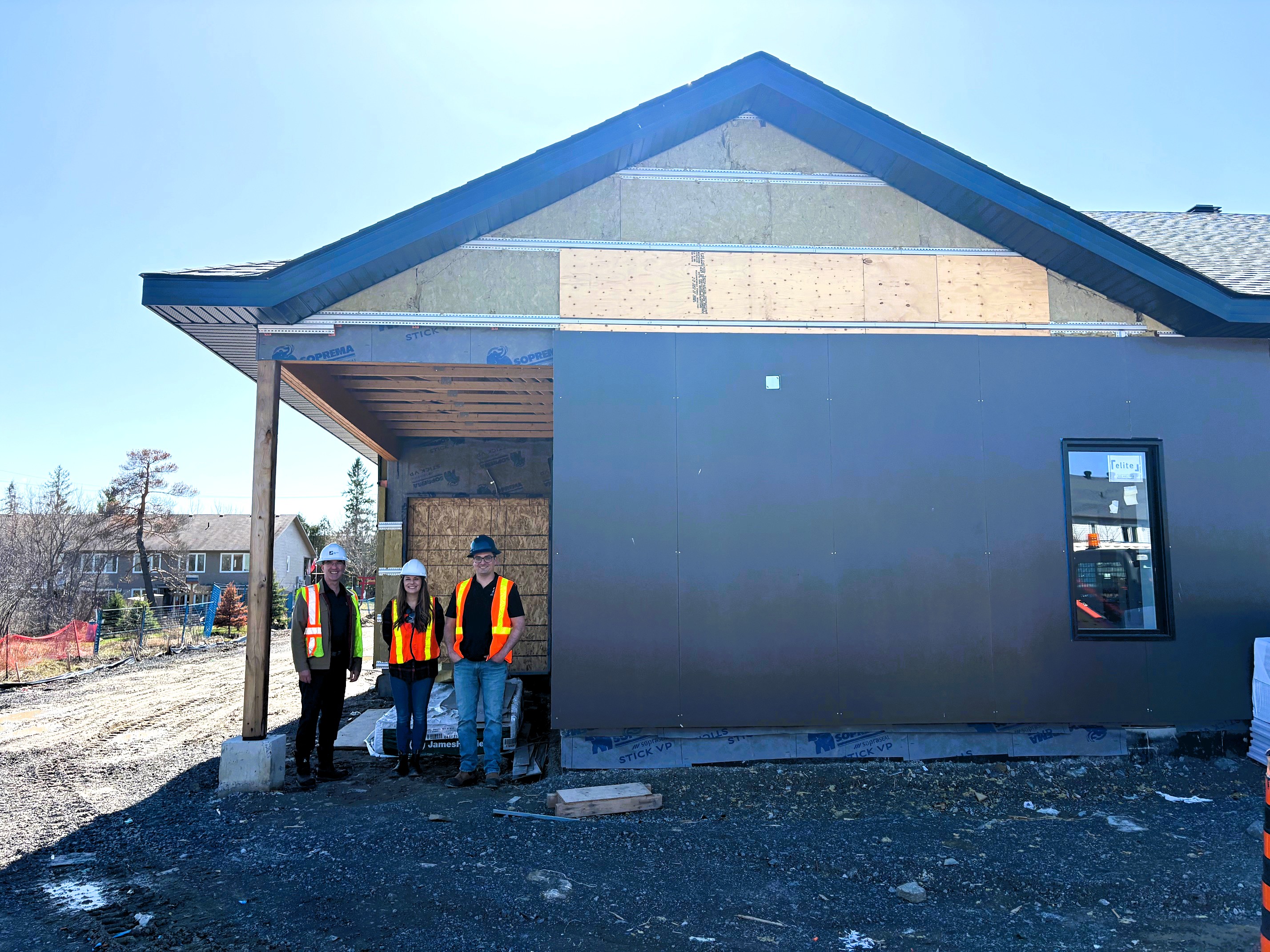 Two men and a woman in orange and yellow construction vests and hard hats stand underneath the entrance to a building under construction.