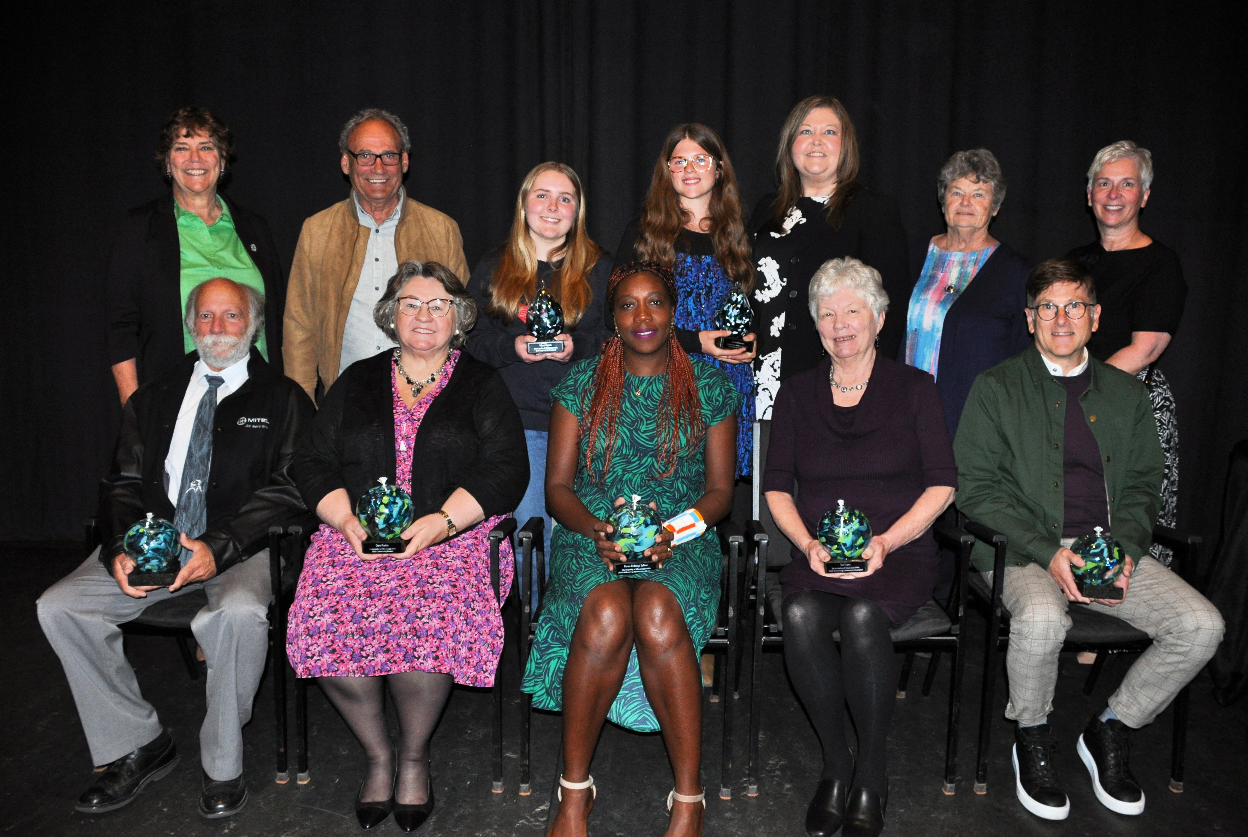 People gather pose for a photo in two rows. Back row is standing, front row is seated with seven holding awards.