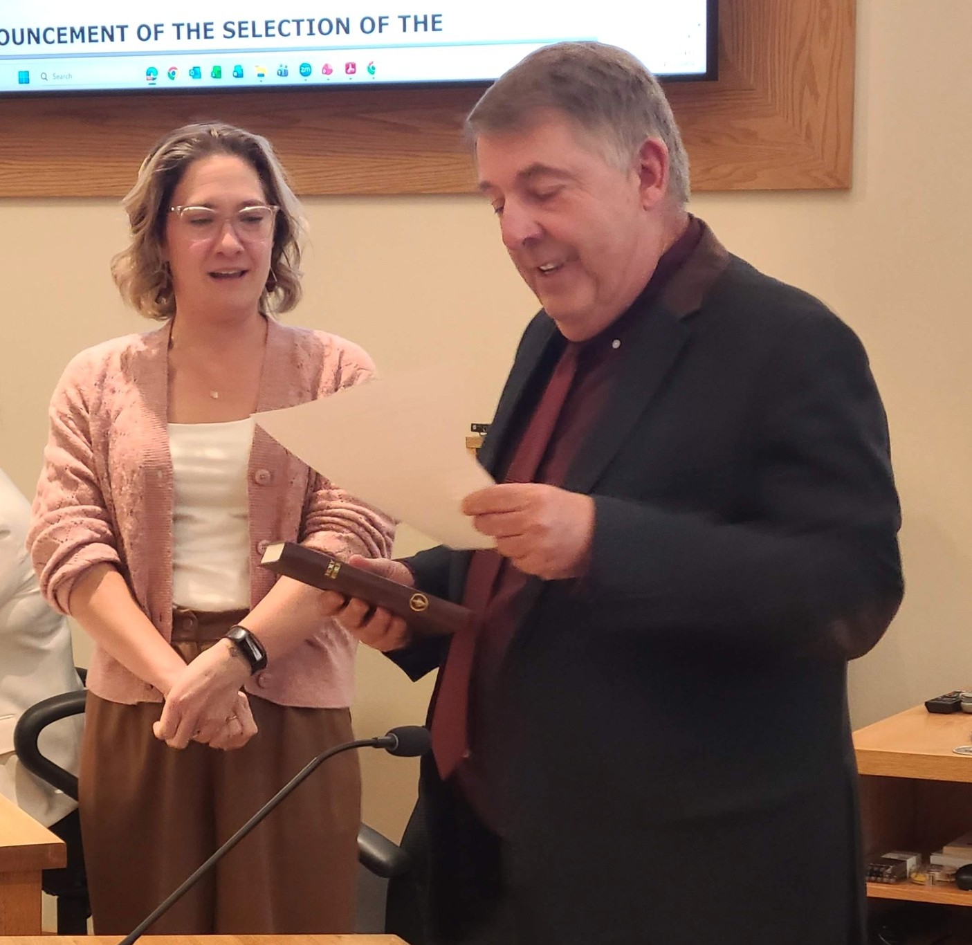 Man in suit reads from a piece of paper while woman in glasses looks on in the background