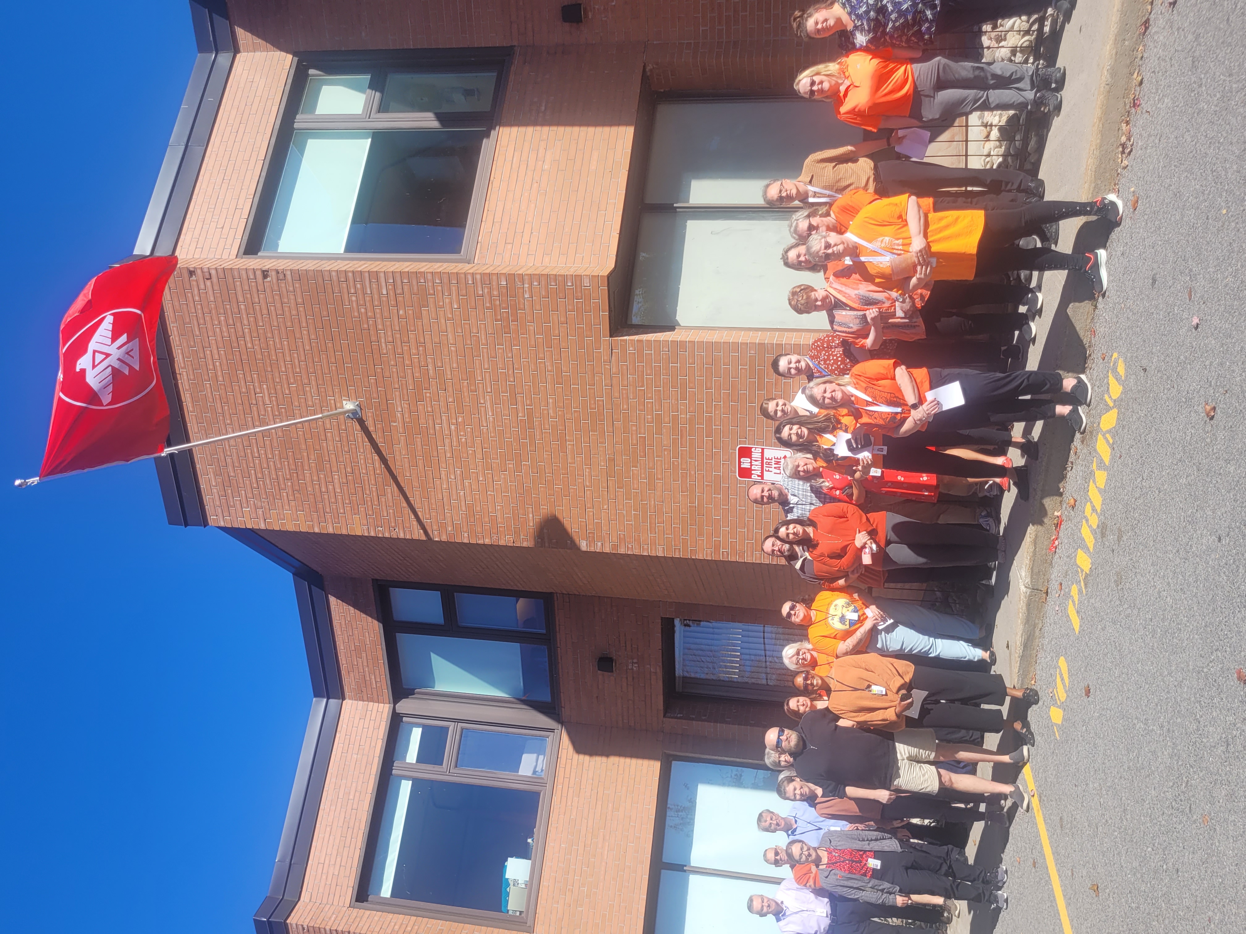 A group of people, mostly wearing orange shirts, stand in front of a brick building with an Indigenous flag flying above