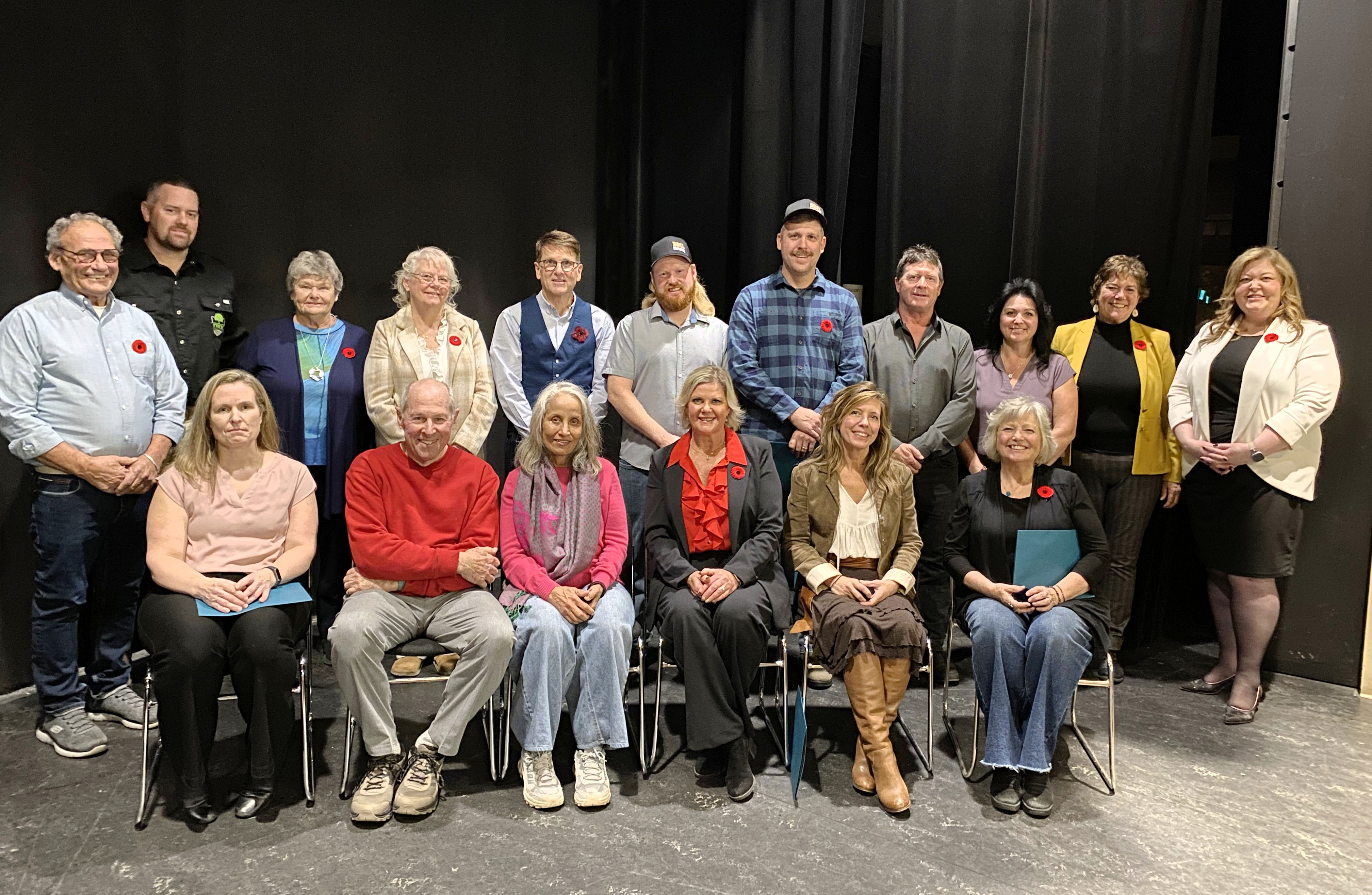 Two rows of people pose for a photo on a stage against a black curtain. Back row is standing, front row is seated.